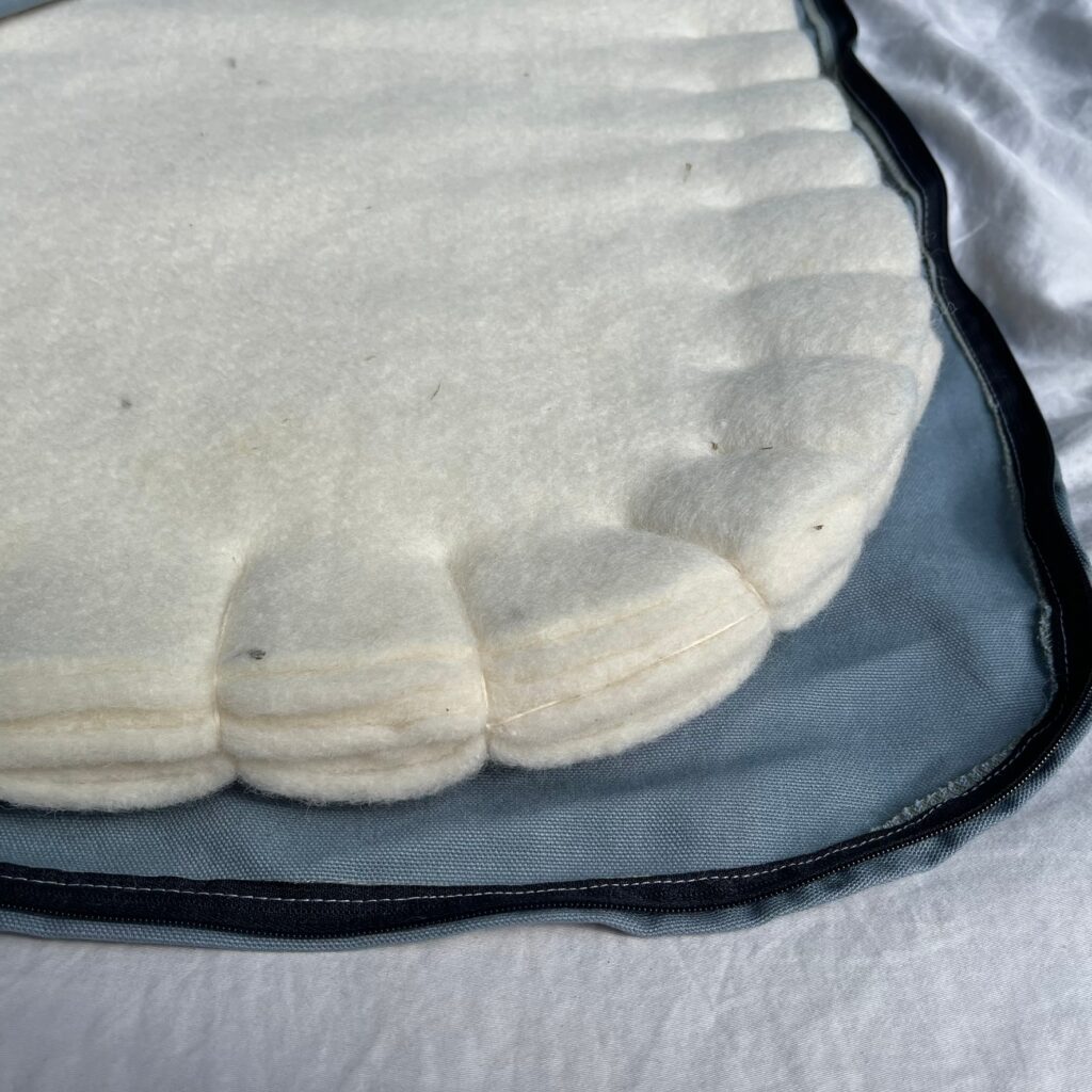 Corner of a wool sleeping pad with the cotton cover unzipped, showing three stacked layers of cream wool felt with scalloped edges