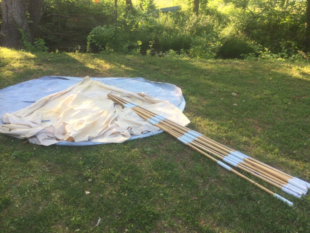 Canvas tipi laid out loosely on a round blue ground cloth on a lawn, with a bundle of jointed wooden poles fitted with pale sleeves lying across it