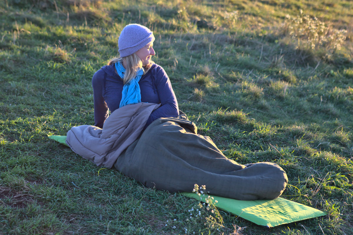 Woman in a beanie sitting up in an olive wool sleeping bag on a pad on an open grassy bald in evening light