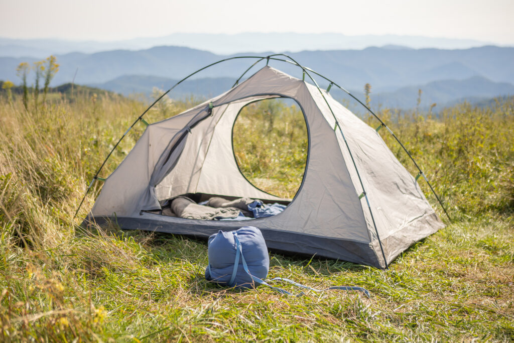 Blue Rewilder wool sleeping bag compressed with straps on the grass in front of a small grey dome tent on a mountain bald, blue ridgelines fading into the distance