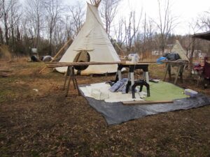 Tipi site with poles laid across sawhorses and a work table on a tarp beside the tipi, bare trees behind