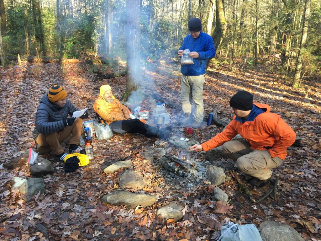 Four campers in puffy jackets and hats around a smoky campfire in a leaf-covered winter hardwood forest