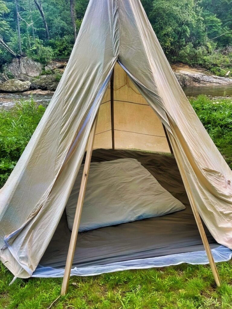 Open door of a canvas tipi tent beside a mountain creek, with a grey wool sleeping pad on a ground cloth inside