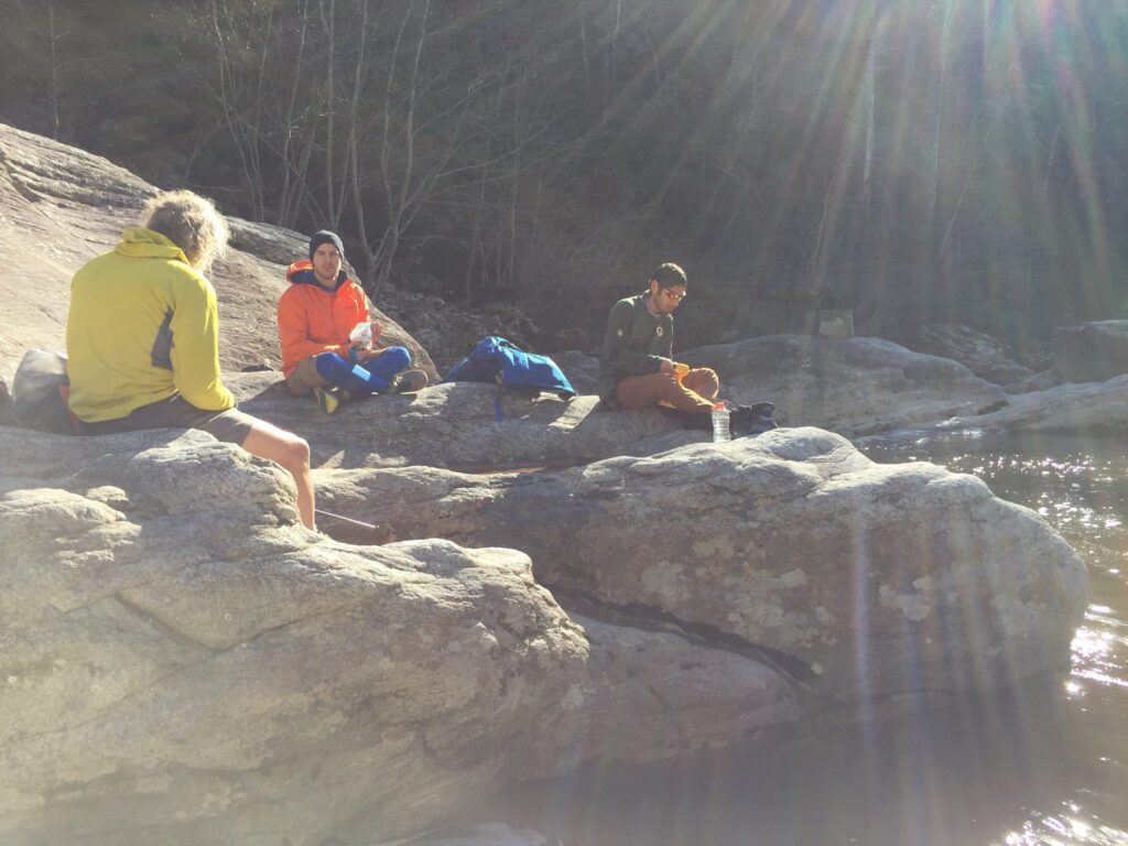 Three hikers resting on sunlit river boulders in a leafless winter gorge, one in a bright synthetic jacket and one in a wool sweater, packs set down beside them