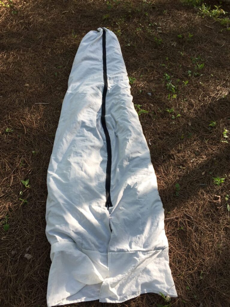 White tapered sleep sack with a full-length black zipper down the centre, laid on pine needles under dappled shade