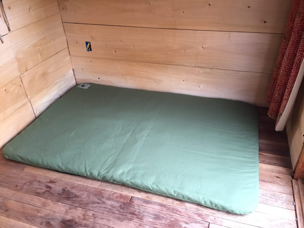 Green wool felt sleeping pad used as a full floor bed on a hardwood floor in a small pine-panelled cabin