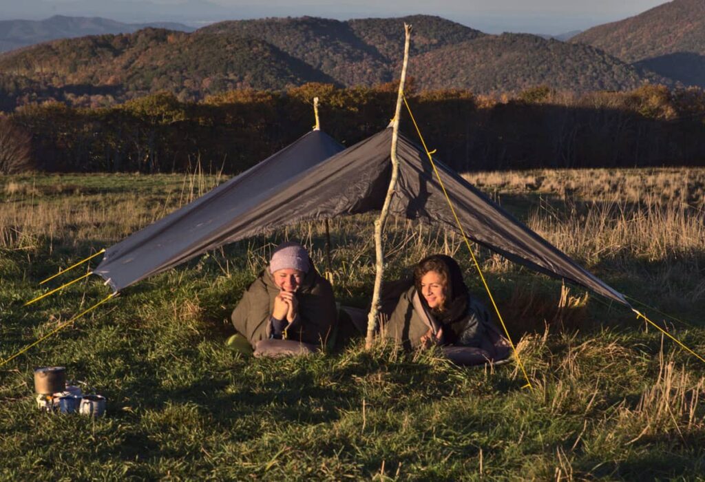 Two women smiling from inside olive wool sleeping bags under a grey tarp pitched on a bare stick on a mountain bald in autumn, wooded ridges and blue mountains behind