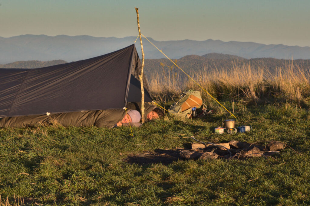 Two people asleep in olive wool sleeping bags under a dark tarp pitched on a single forked stick on a grassy mountain bald at sunrise, with a stove, pack and blue mountain ridges behind