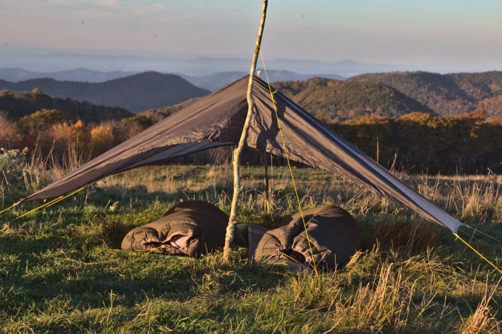 Two sleepers zipped completely into wool sleeping bags, heads inside, under a low tarp on a grassy bald