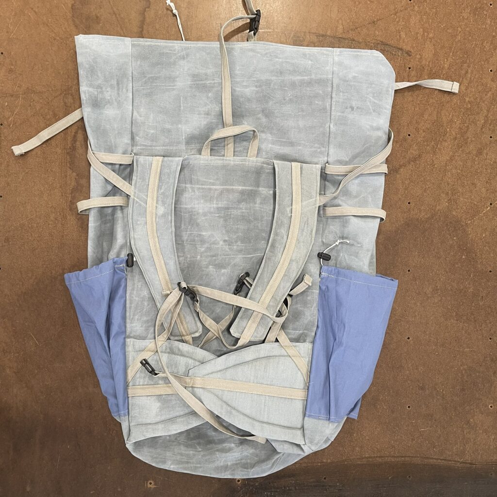 Back of the 65 L waxed canvas backpack laid flat: padded shoulder straps, sternum strap, padded hip belt, blue side pockets and webbing adjusters