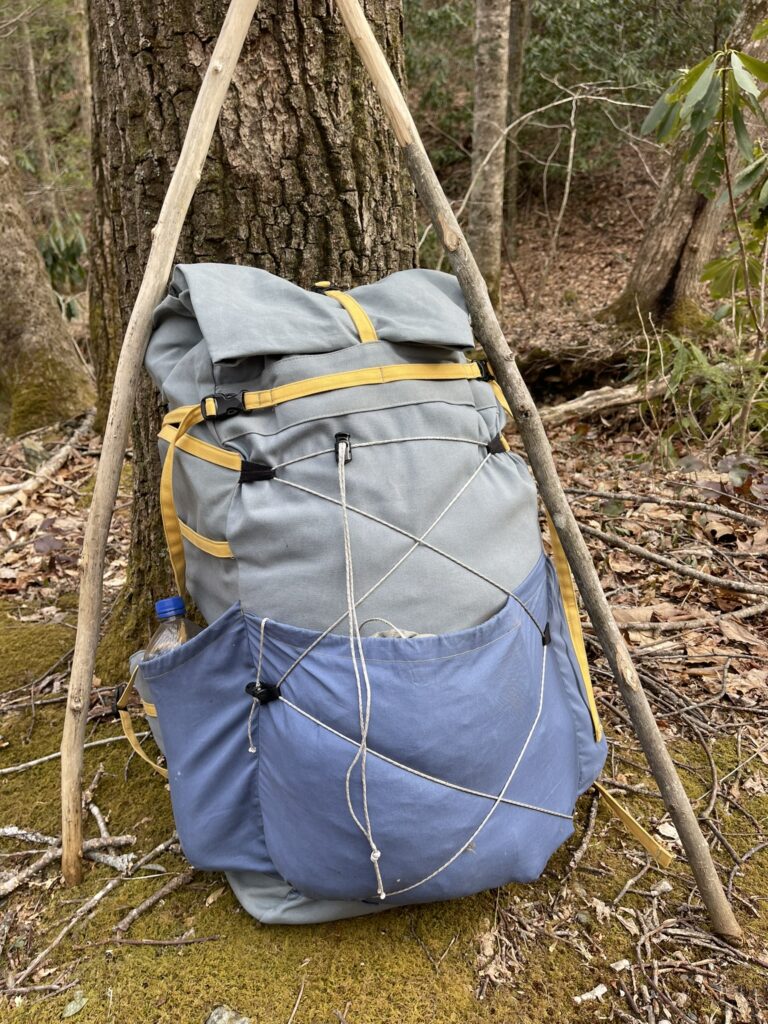Grey organic canvas 65-litre backpack with blue pockets and yellow straps leaning against an oak trunk between two wooden walking sticks in a hardwood forest