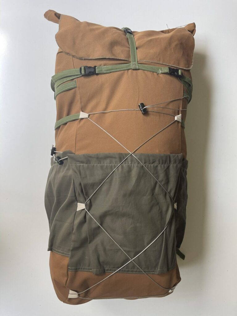 Front view of a tan organic canvas 65-litre backpack with an olive front pocket, green webbing straps and crossed cord lacing, on a white background