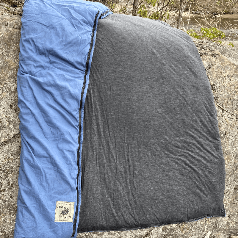 Aurora Borealis wool sleeping bag unzipped and opened flat on a rock by a river, blue cotton shell on one side and charcoal merino liner on the other