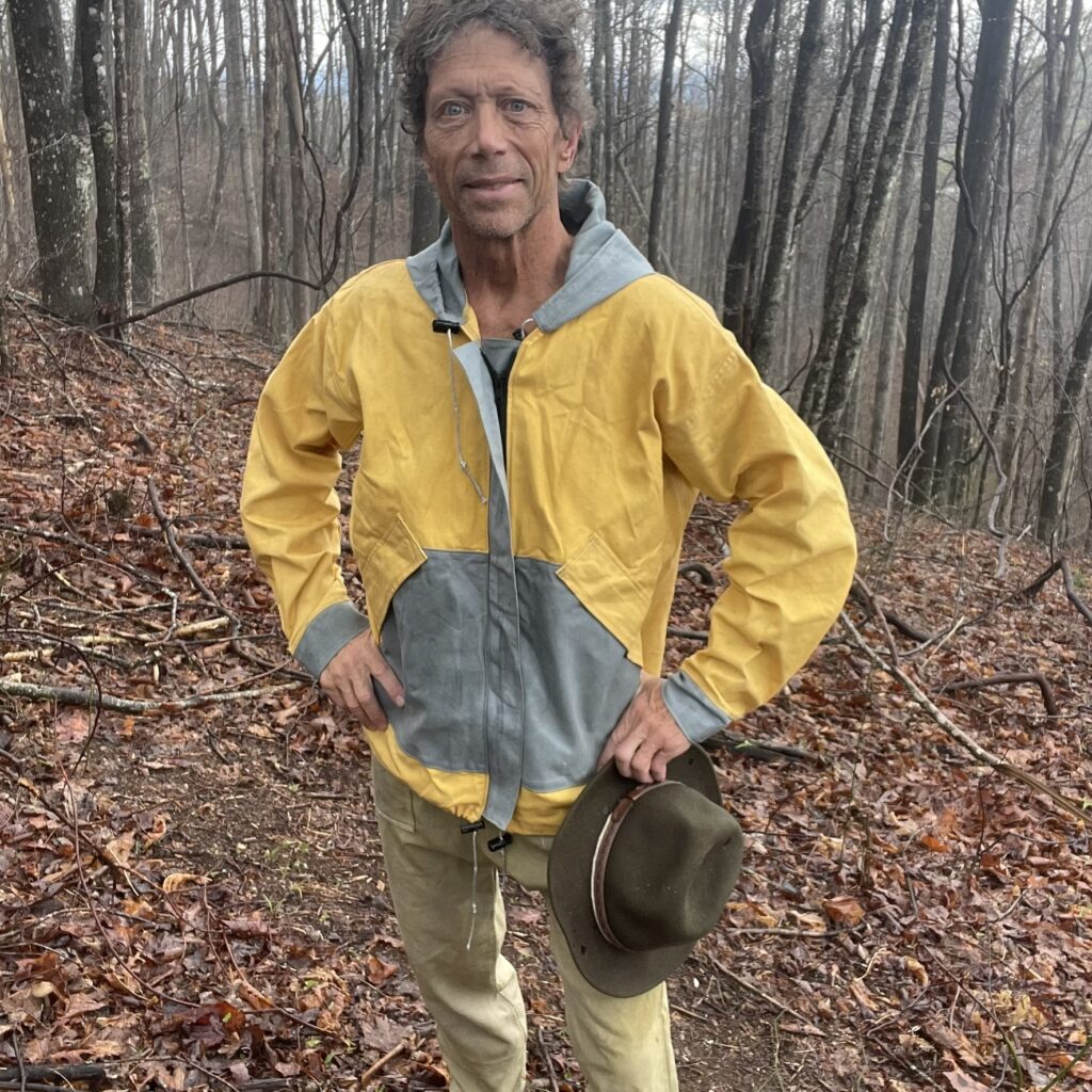 Patrick Clark in a yellow and grey waxed canvas rain jacket, hat in hand, in a bare winter forest
