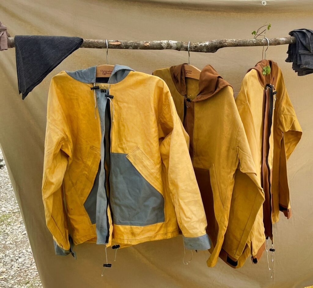 Three waxed canvas rain jackets in yellow, tan and gold hanging on a wooden pole in front of a canvas tent wall, wool layers draped beside them
