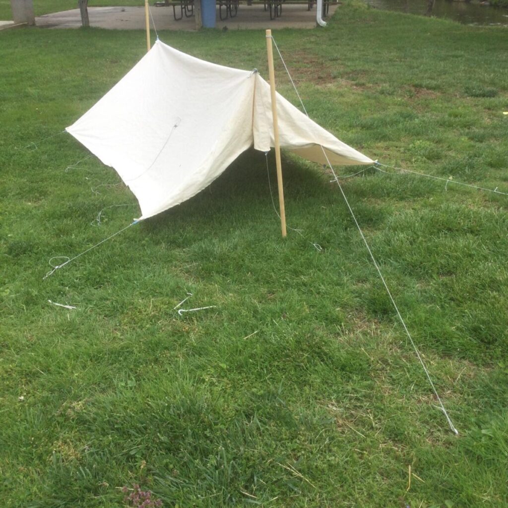 Canvas tarp pitched as a lean-to with one edge raised on two wooden poles and the back edge staked low, guylines running out to stakes on grass