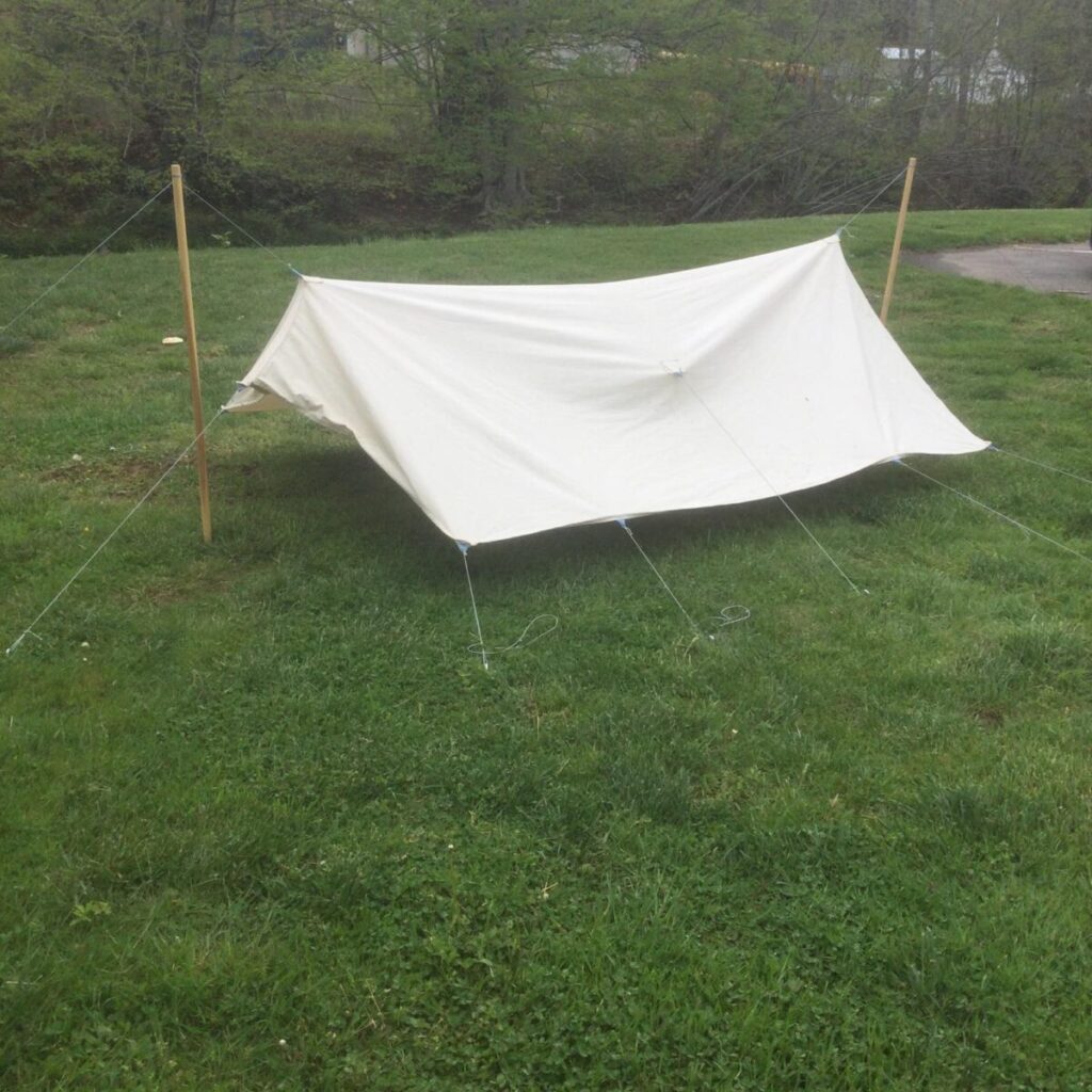 White organic canvas tarp pitched as an A-frame between two upright wooden poles on a lawn, corners and mid-panel tie-outs guyed to stakes