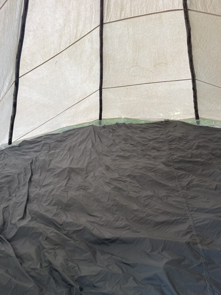 Inside a canvas tipi looking at the cream canvas wall and three dark wooden poles, with a grey ground cloth covering the floor