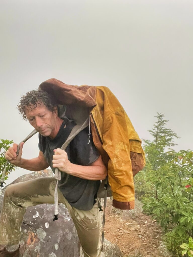 Hiker with wet hair hoisting a canvas backpack onto his shoulders in thick fog on a rocky summit, an ochre waxed canvas jacket draped over the pack
