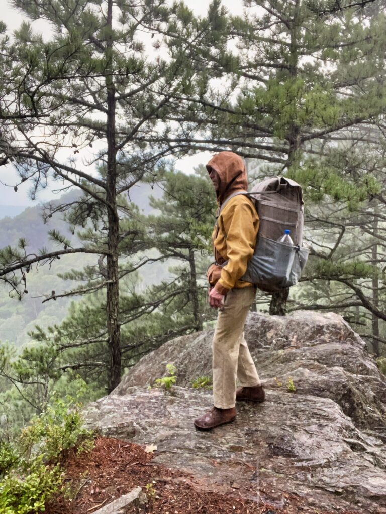 Hiker in a hooded waxed canvas jacket standing on a wet boulder in the rain with the 65 L canvas backpack, a water bottle in its side pocket, misty forested valley behind