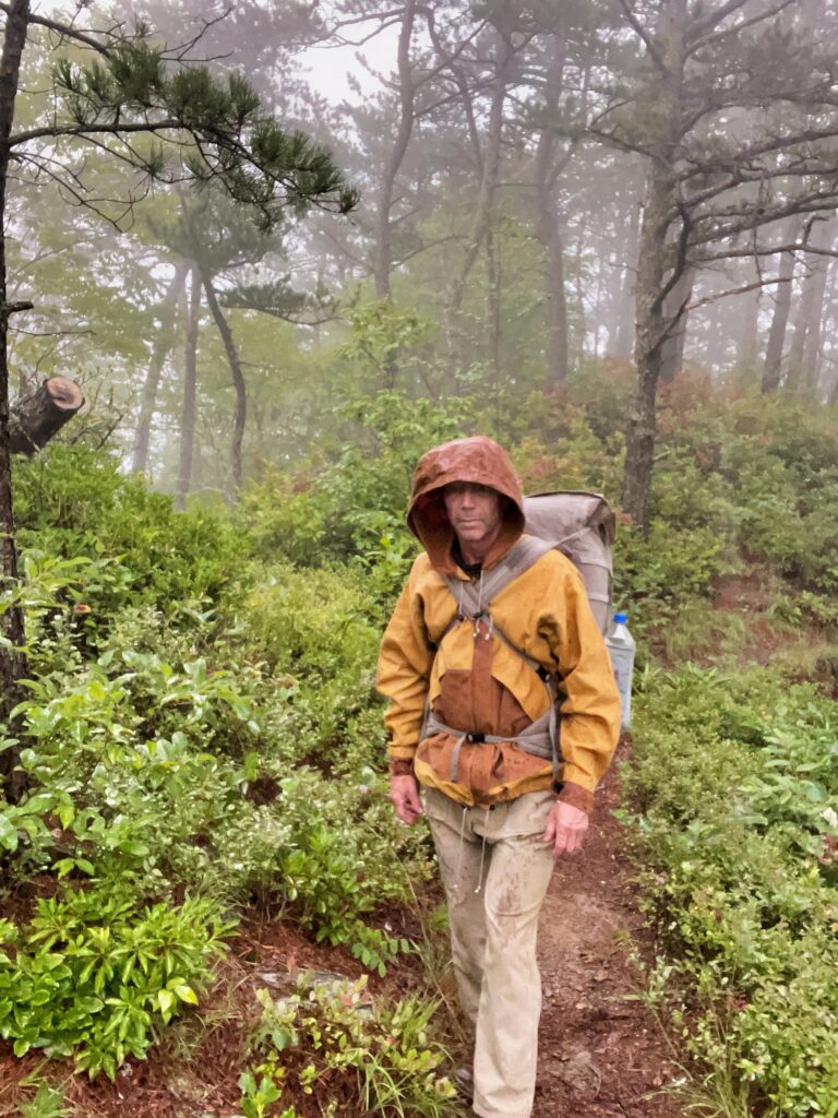 Hiker in a hooded waxed canvas rain jacket and 65 L canvas backpack walking a foggy trail through wet pines in steady rain